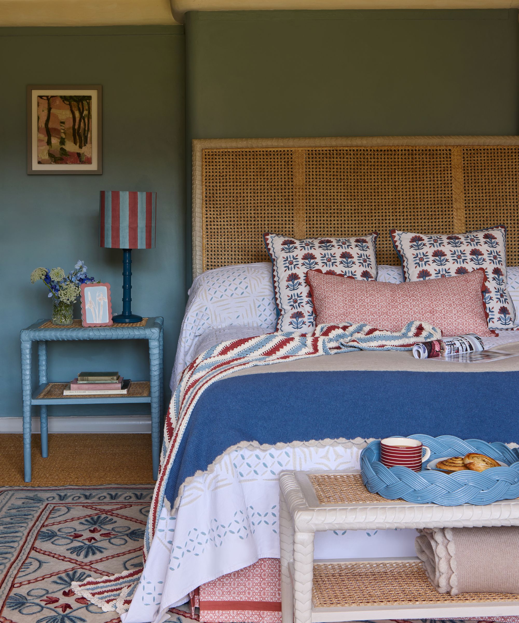 blue bedroom by birdie fortescue with a rattan headboard, white bed linen, floral blue and red pillows, blue side table, red and blue striped lampshade