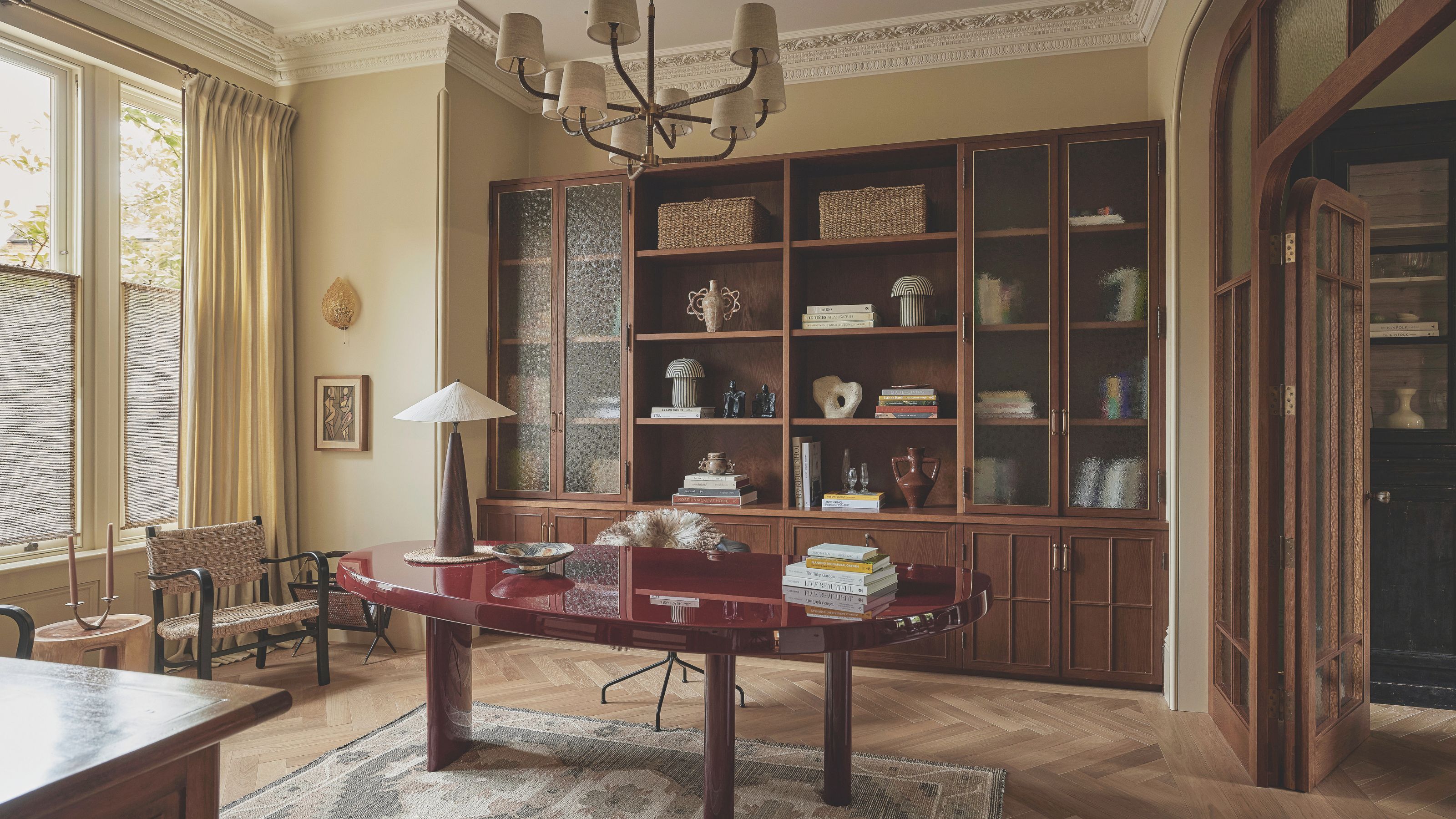 A large study with a wall of glass fronted, built in wooden cabinets and an oval glossy red desk in the middle of the room