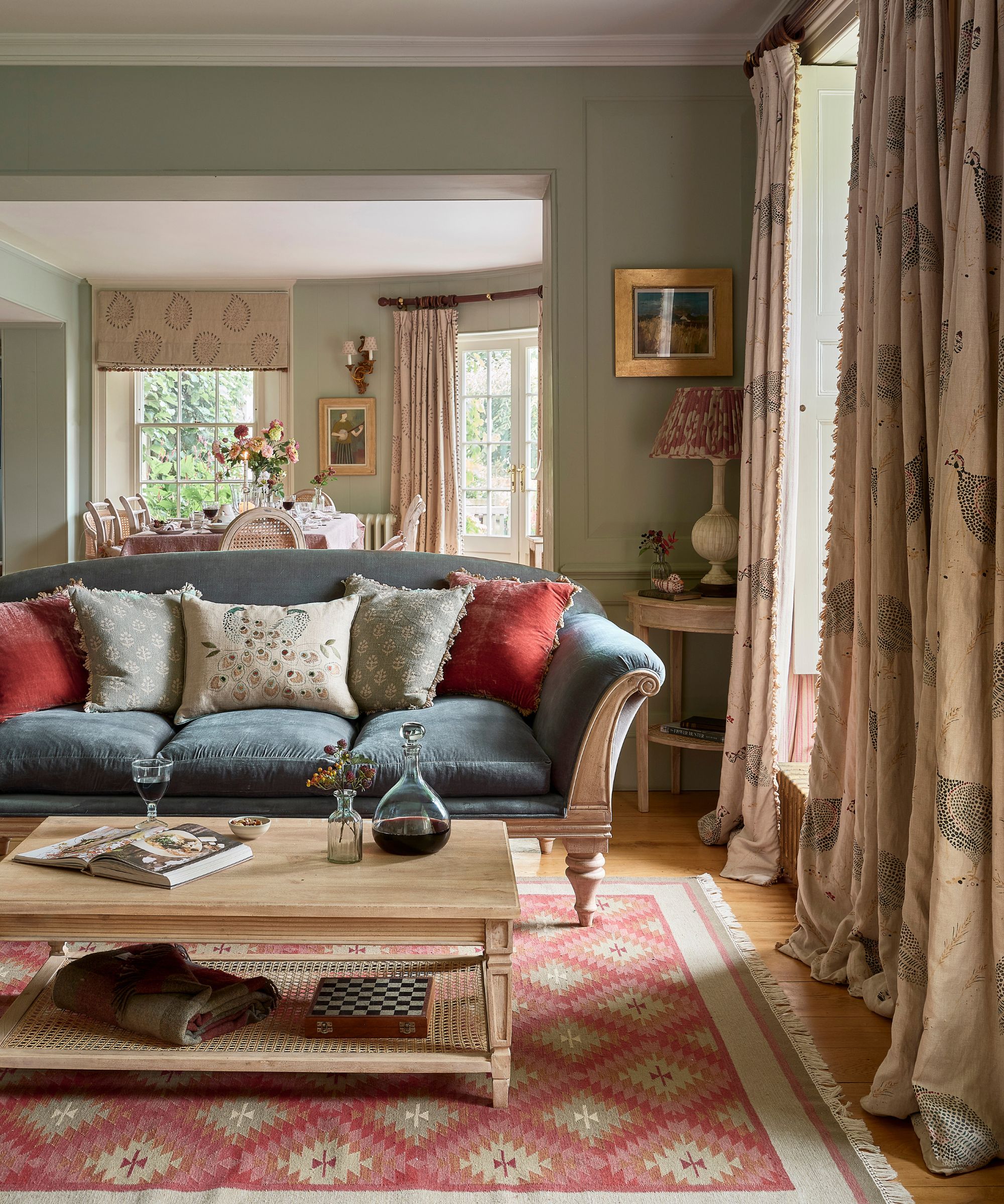 A sitting room with a dark teal velvet Georgian style sofa with five patterned scatter cushions, a pale wood coffee table with a decanter of wine and a wine glass, a kilim rug in bright pink and heavy long curtains with a guinea fowl print