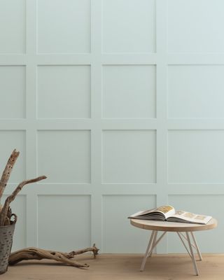 A paneled wall painted in Benjamin Moore's Woodlawn Blue HC-147 next to wood flooring and a small round table with an open book on it and a basket with large twigs and one on the floor