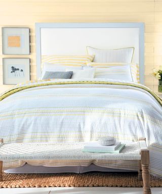 A bedroom with a muted yellow painted wall, a light blue bed frame, light blue and yellow striped bedding and throw pillows, two framed wall art prints, and a brown jute rug underneath it