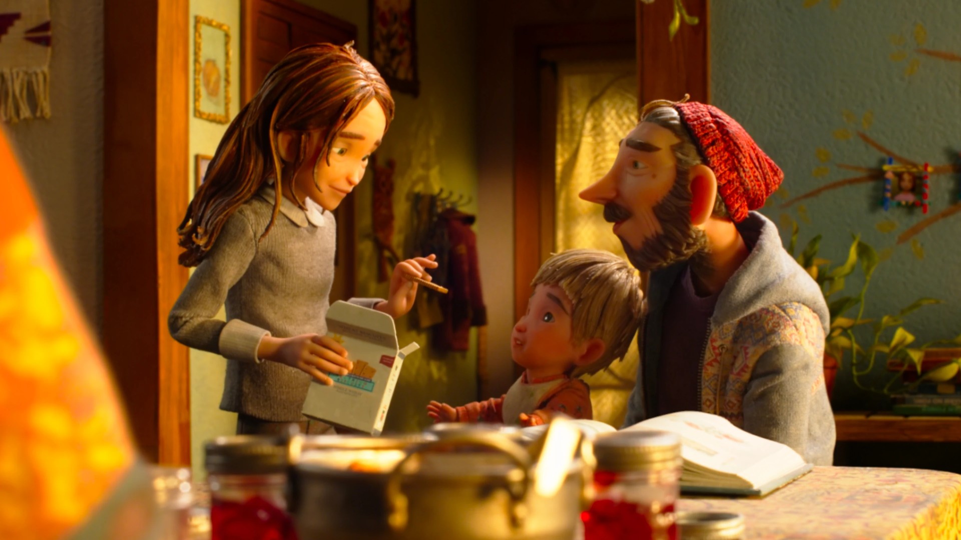 A stop-motion still showing a teenage girl holding a cracker box, a toddler and a bearded man in a red beanie gathered around a kitchen table lit by warm lamplight.