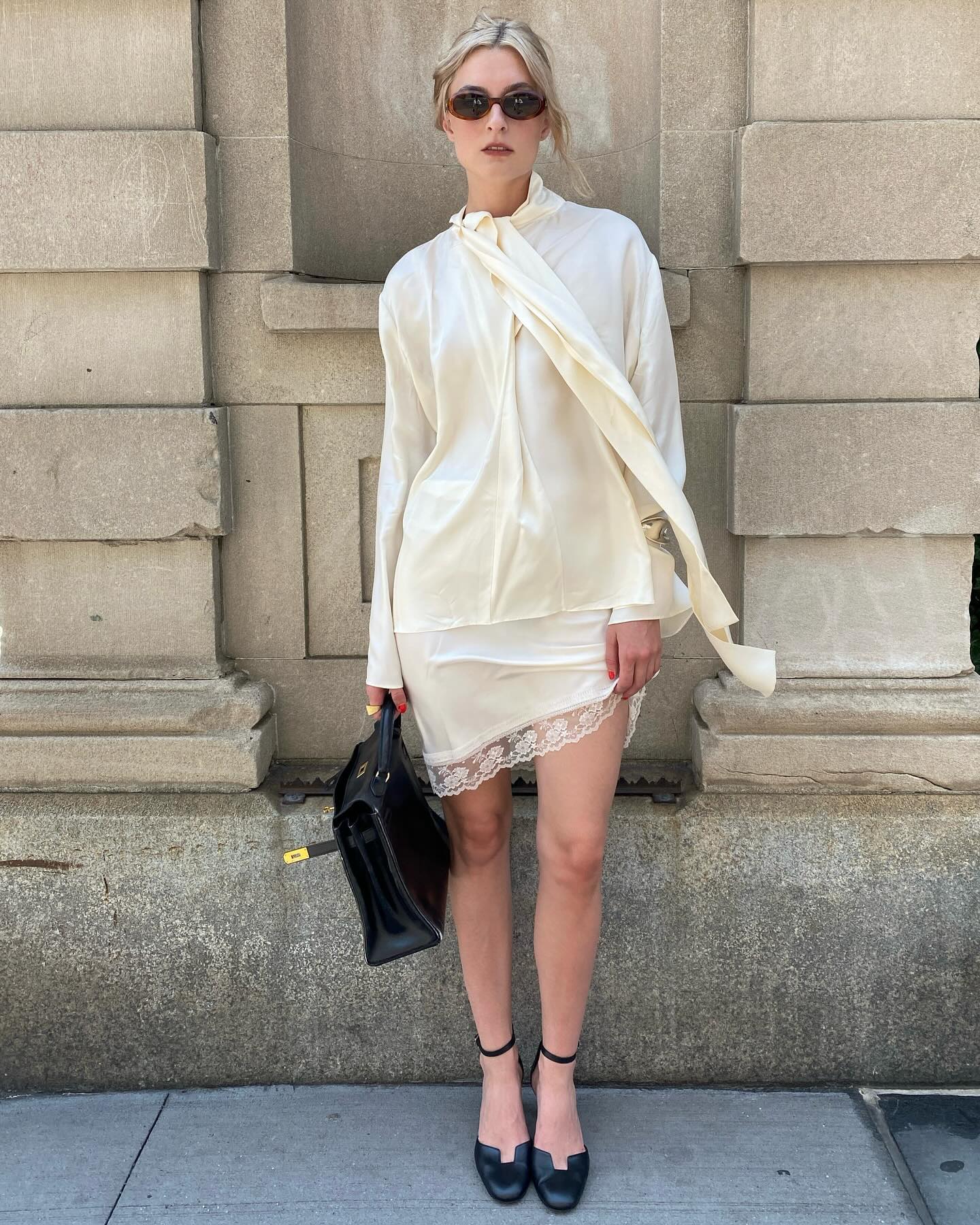 Christie Tyler wearing a cream satin blouse and lace-trim skirt with black heels and sunglasses.