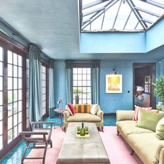 orangery with blue walls brown framed windows and roof light and pink rug with green velvet sofas