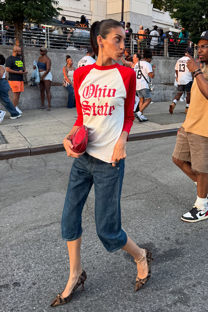 A woman wearing a red baseball tee and denim capri pants.