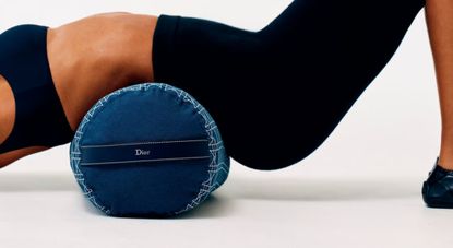 A woman dressed in a fitness matching set of cropped top and leggings reclines on a padded back cushion with a checkered motif in black and white and the logo "Dior".