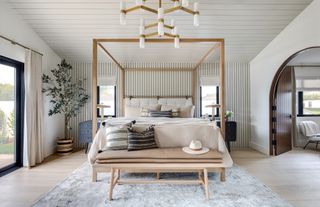 white bedroom with shiplap walls and ceiling, modern pendant, blond wood four poster and bench, pale wood flooring, gray rug, black side tables, plant, stripe cushions