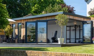 Timber-clad garden gym with folding glass doors open, showing squat rack and bench, olive tree on deck.