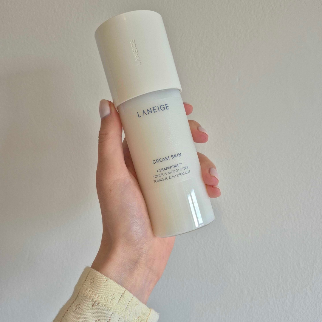 A close-up of Digital Beauty Writer, Naomi Jamieson's hand holding the LANEIGE Cream Skin Cerapeptide Toner and Moisturiser, in front of a cream wall