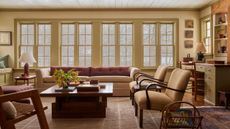 Warm living room with large butter yellow windows, a large beige sofa with pink cushions, two midcentury armchairs, a wooden coffee table, a striped rug, and a lamp in the corner.
