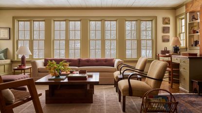Warm living room with large butter yellow windows, a large beige sofa with pink cushions, two midcentury armchairs, a wooden coffee table, a striped rug, and a lamp in the corner.