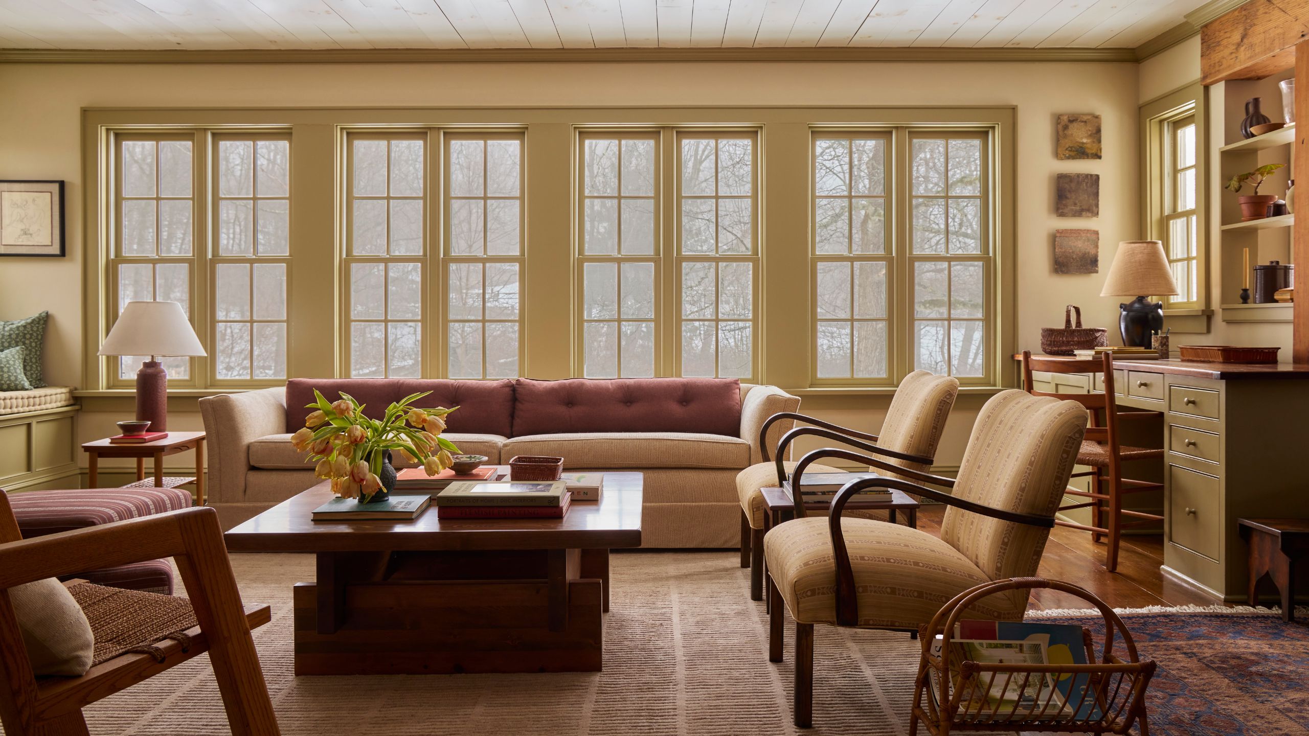Warm living room with large butter yellow windows, a large beige sofa with pink cushions, two midcentury armchairs, a wooden coffee table, a striped rug, and a lamp in the corner.
