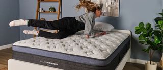 The image shows a light haired woman smiling as she dives onto a mattress that is placed in a pale blue bedromm