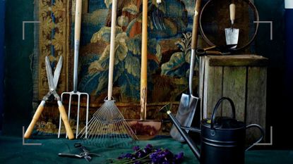 Dark room with tapestry wall hanging with garden tools in front with a watering can and wooden crate to highlight January garden jobs