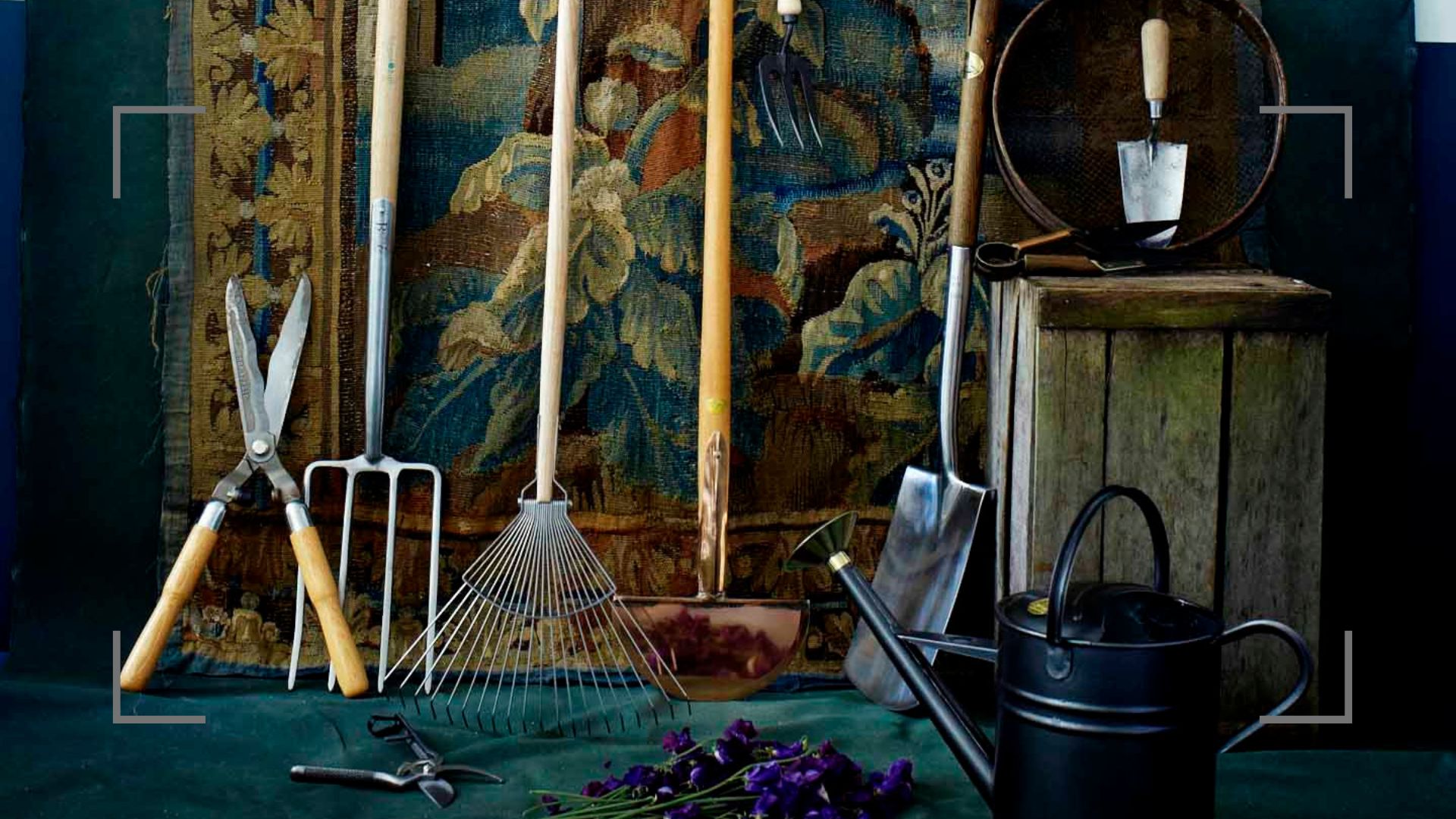 Dark room with tapestry wall hanging with garden tools in front with a watering can and wooden crate to highlight January garden jobs