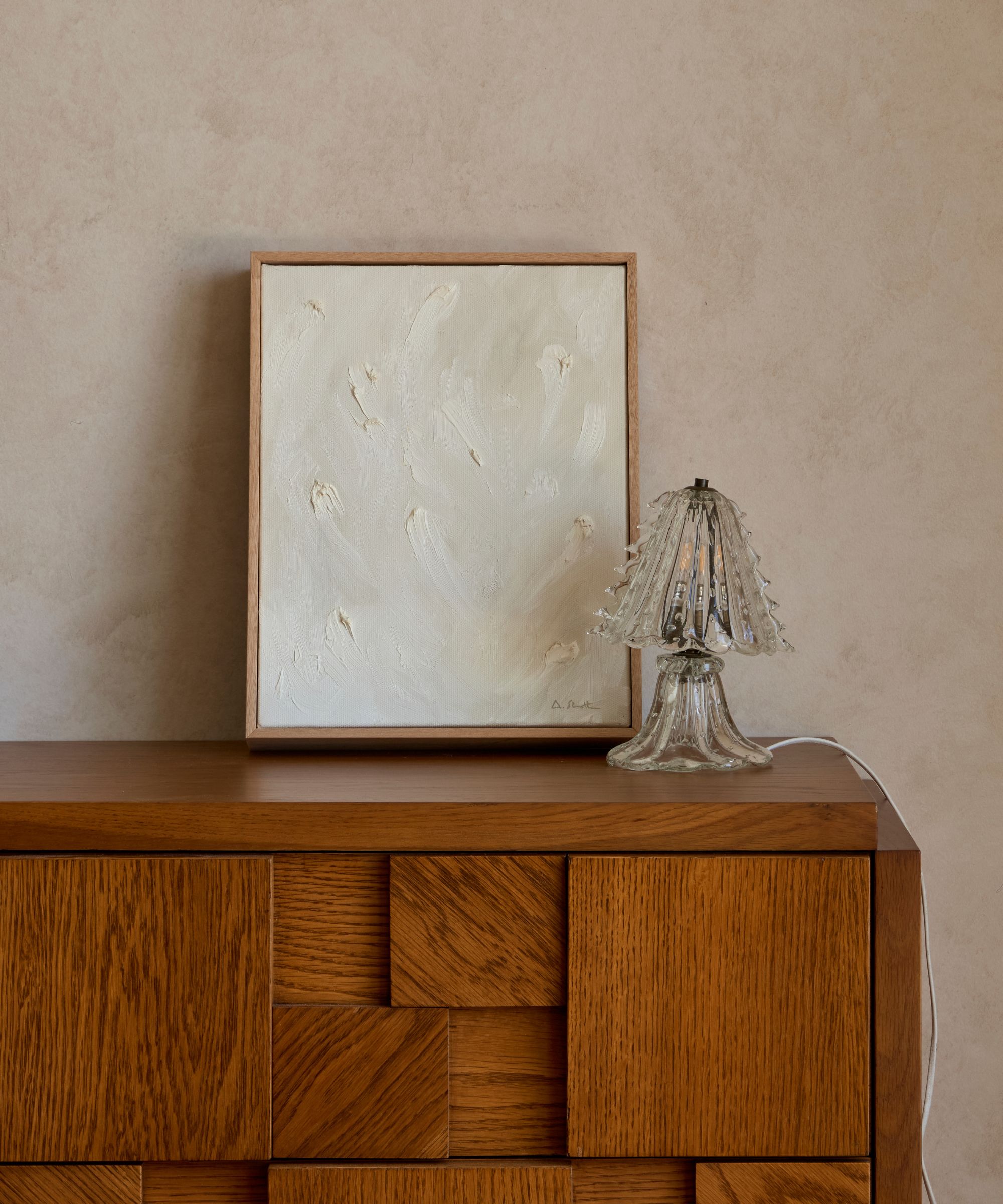 1940s-style Murano glass table lamp with a fluted shade and sculptural base, styled beside a textured abstract painting on a mid-century wooden cabinet.