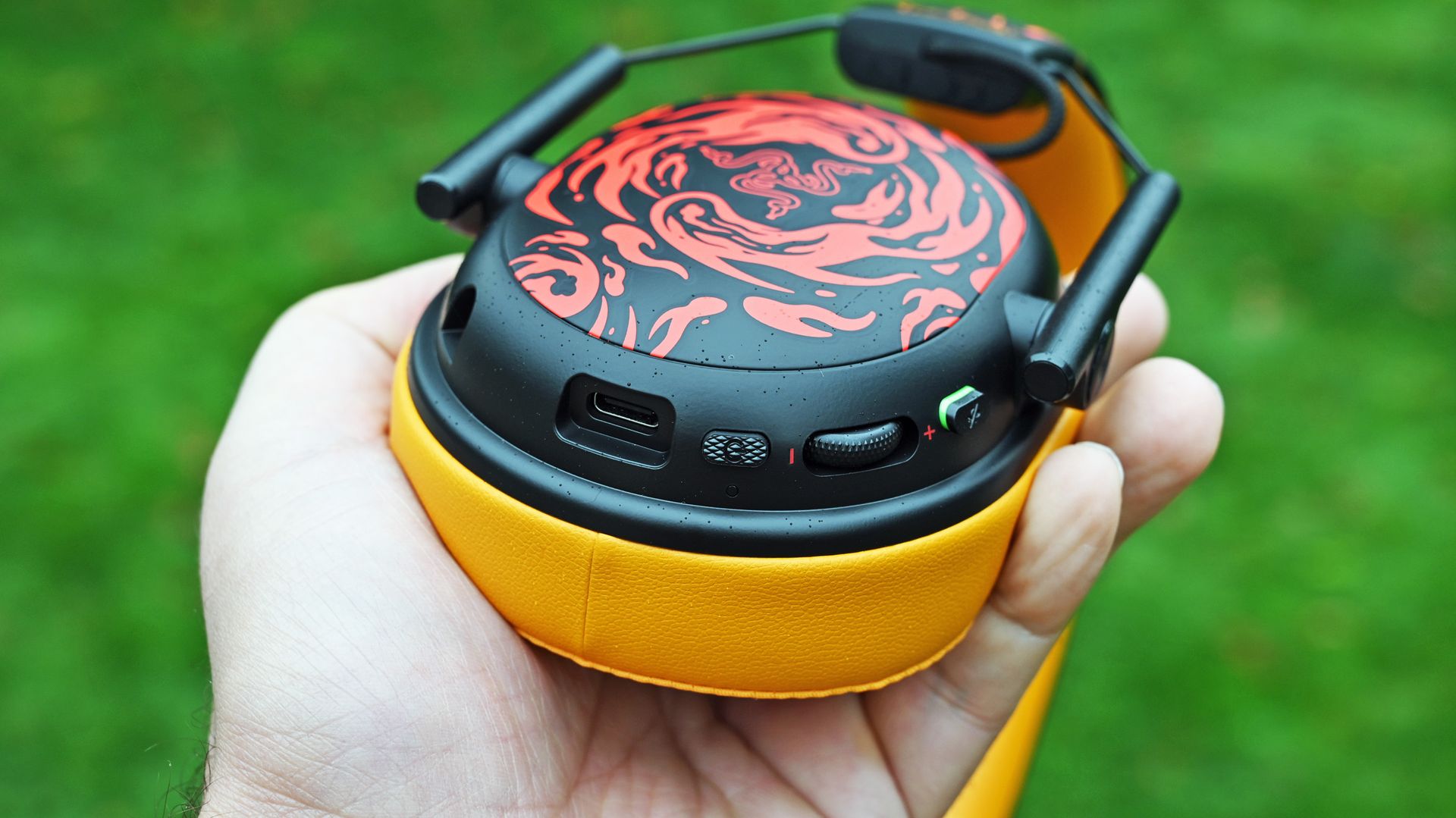 Hand holding the headset earcup, showing bottom controls including a USB-C port, power button, volume dial, and mic mute button against a yellow cushion.