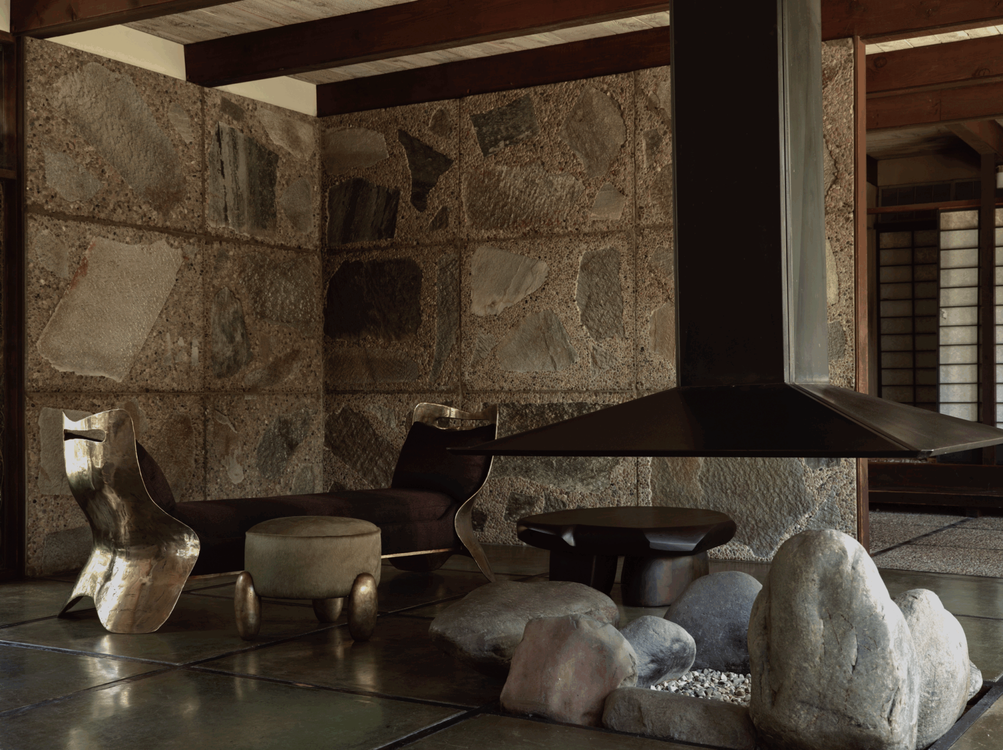 A series of brass-clad sculptural furnishings sits in a brutalist room with rock walls, ornamental stones, and a floating fireplace.