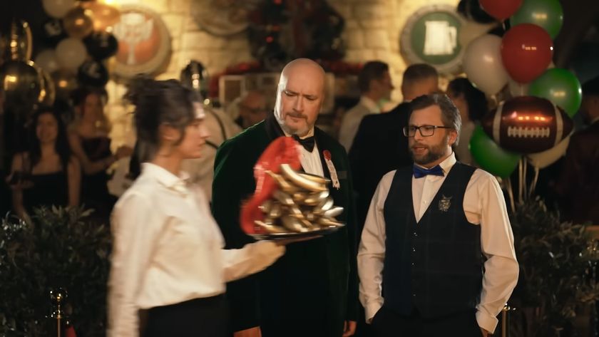 Two men wearing posh clothing at a party watch in confusion as a woman holding a platter of golden bananas with a red plume walks past