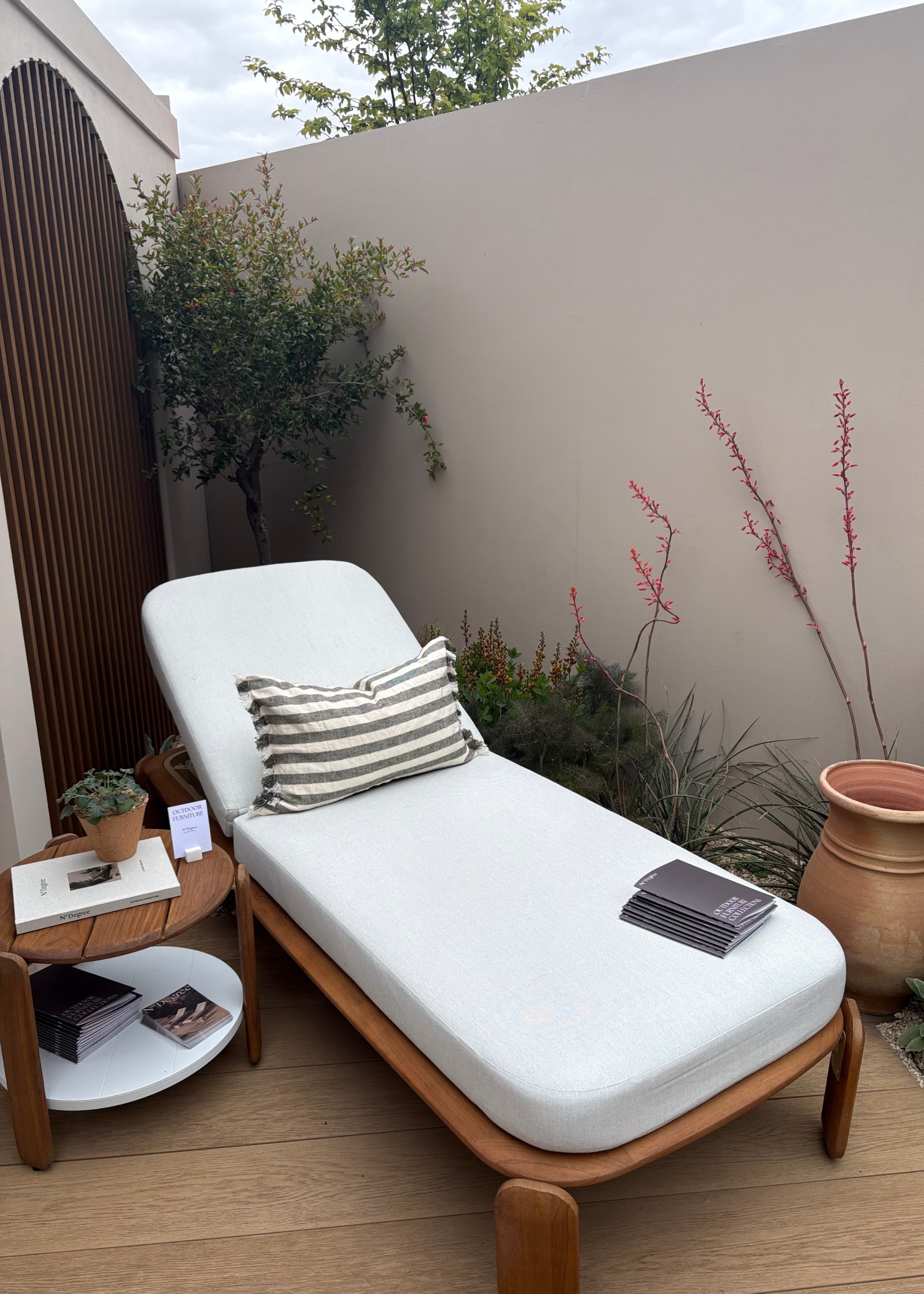 An outdoor space at the Chelsea Flower Show featuring Mediterranean garden design alongside a sun lounger, a wood side table, and a terracotta pot