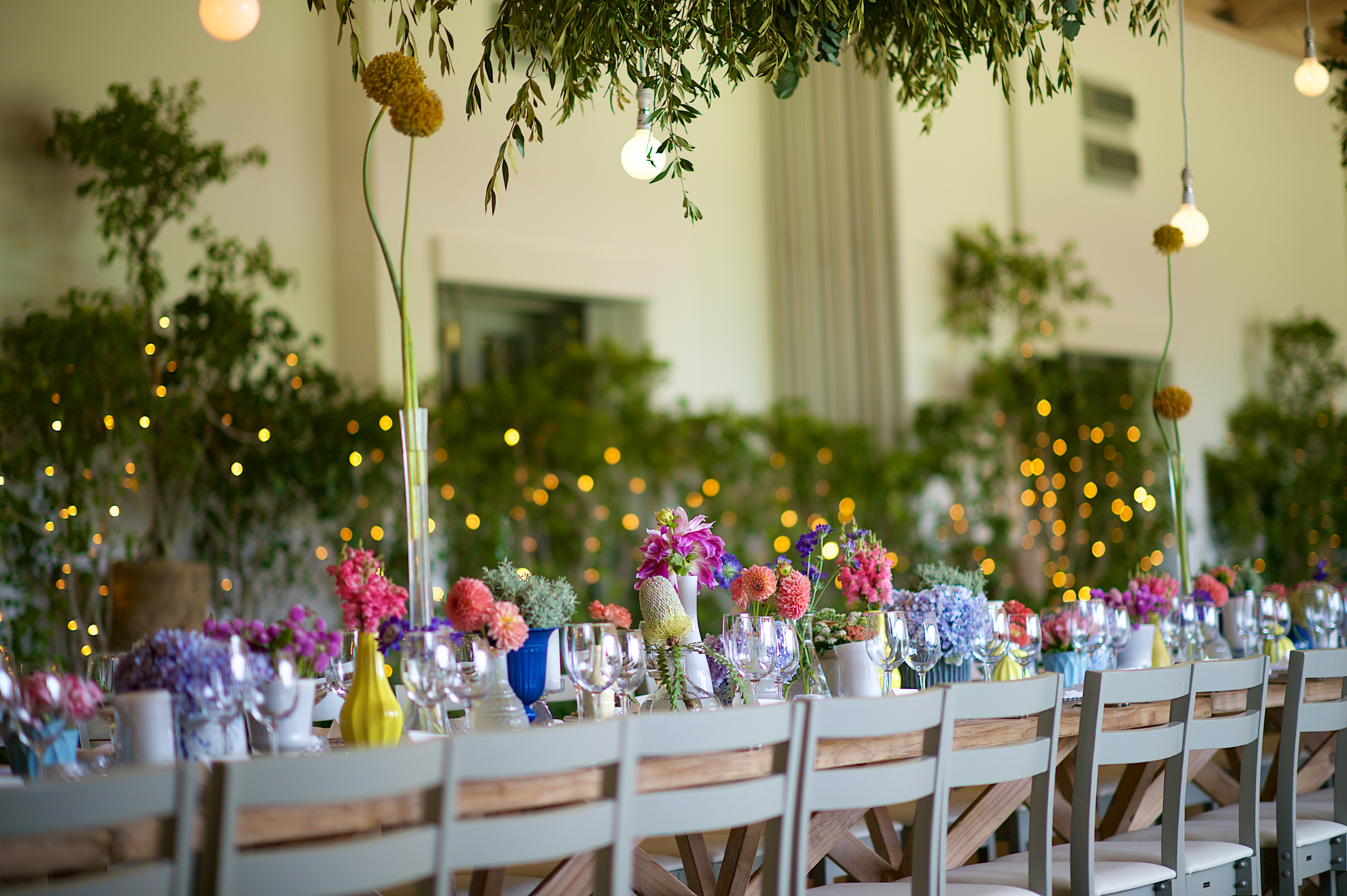 Beautiful Modern Summer Colourful Wedding Flower Arrangements at a dinner party wedding reception. There is hanging LED bulbs and plants hanging from the ceiling. The string fairy lights are defocussed in the background for a pleasant bokeh.