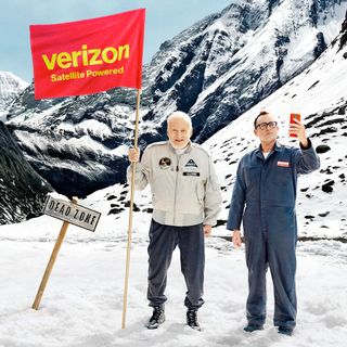 two men, one holding a red and yellow flag and the other a cell phone stand ate base of a snow-covered mountain