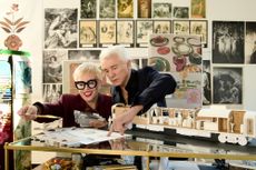 A middle-aged woman dressed in a bordeaux tailored jacket and big, square black glasses and a middle-aged man wearing a black shirt with unbottoned, V neck handle a series of sketches in a studio plastered in photographs and illustrations.