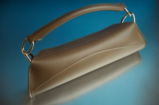 Small brown leather bag features Elongated construction with round stitched top-handle photographed against a metalic blue background