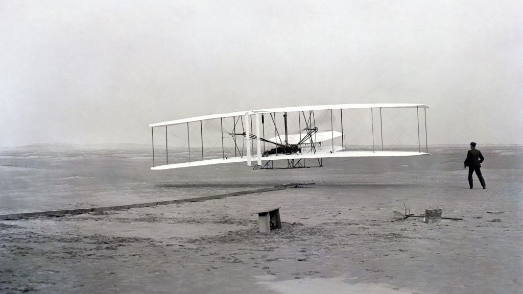 monochrome photograph the Wright Brothers&#039; plane and a man in black standing to the right. 