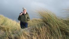 Woman wearing coat and hat walking through reeds on beach against stormy autumnal sky