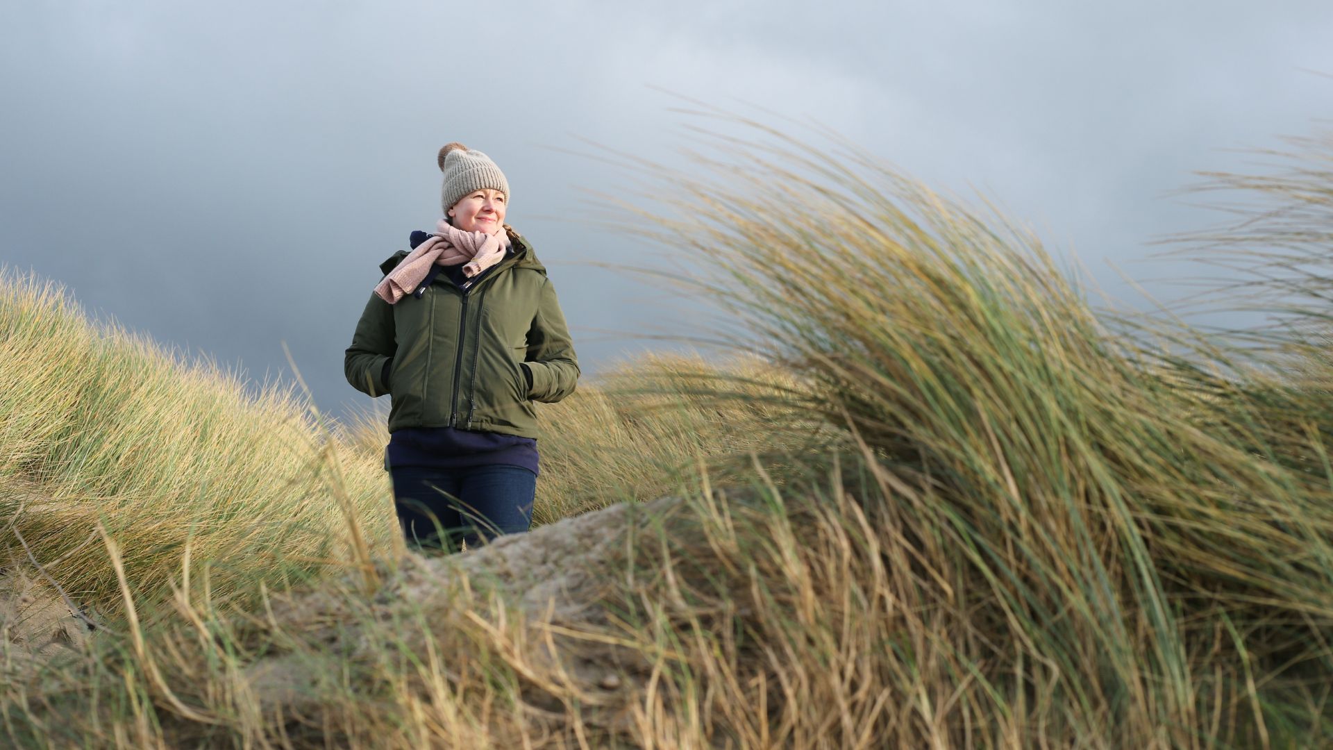 Woman wearing coat and hat walking through reeds on beach against stormy autumnal sky