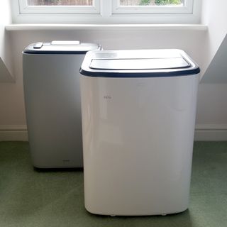 A grey and a white portable air conditioner being tested in a room with green carpet