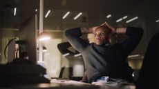 A male cybersecurity professional sitting at a desk in dimly lit room with hands placed on head with a stressed expression.
