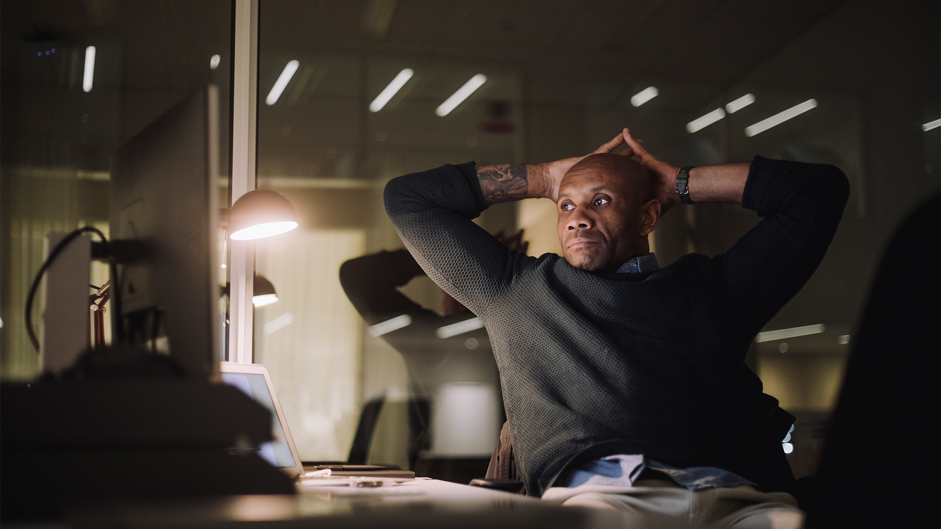 A male cybersecurity professional sitting at a desk in dimly lit room with hands placed on head with a stressed expression.