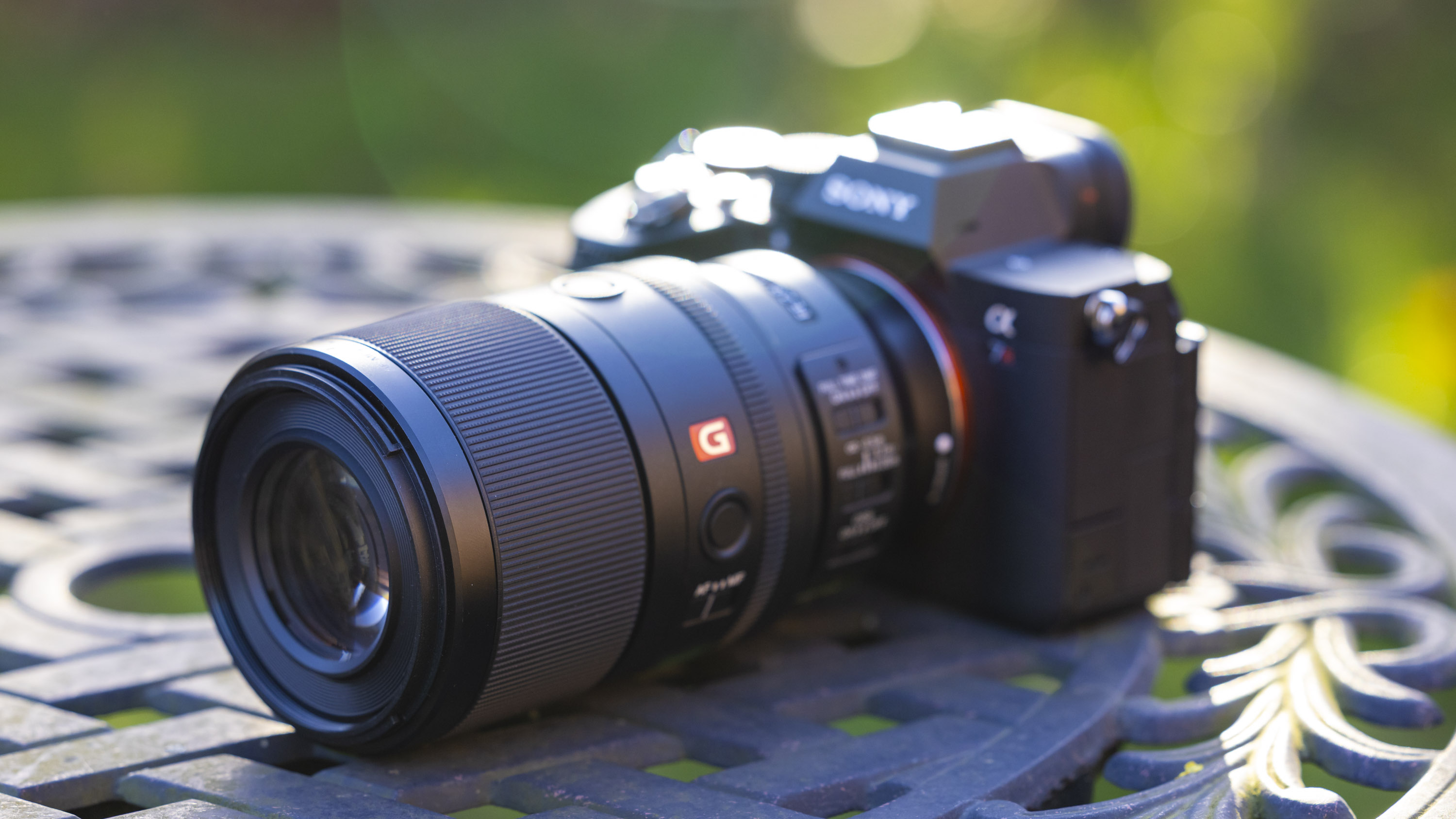 A product shot of the Sony FE 100mm f/2.8 Macro GM OSS attached to a Sony A7R IV outdoors on a metal garden table with a blurry background