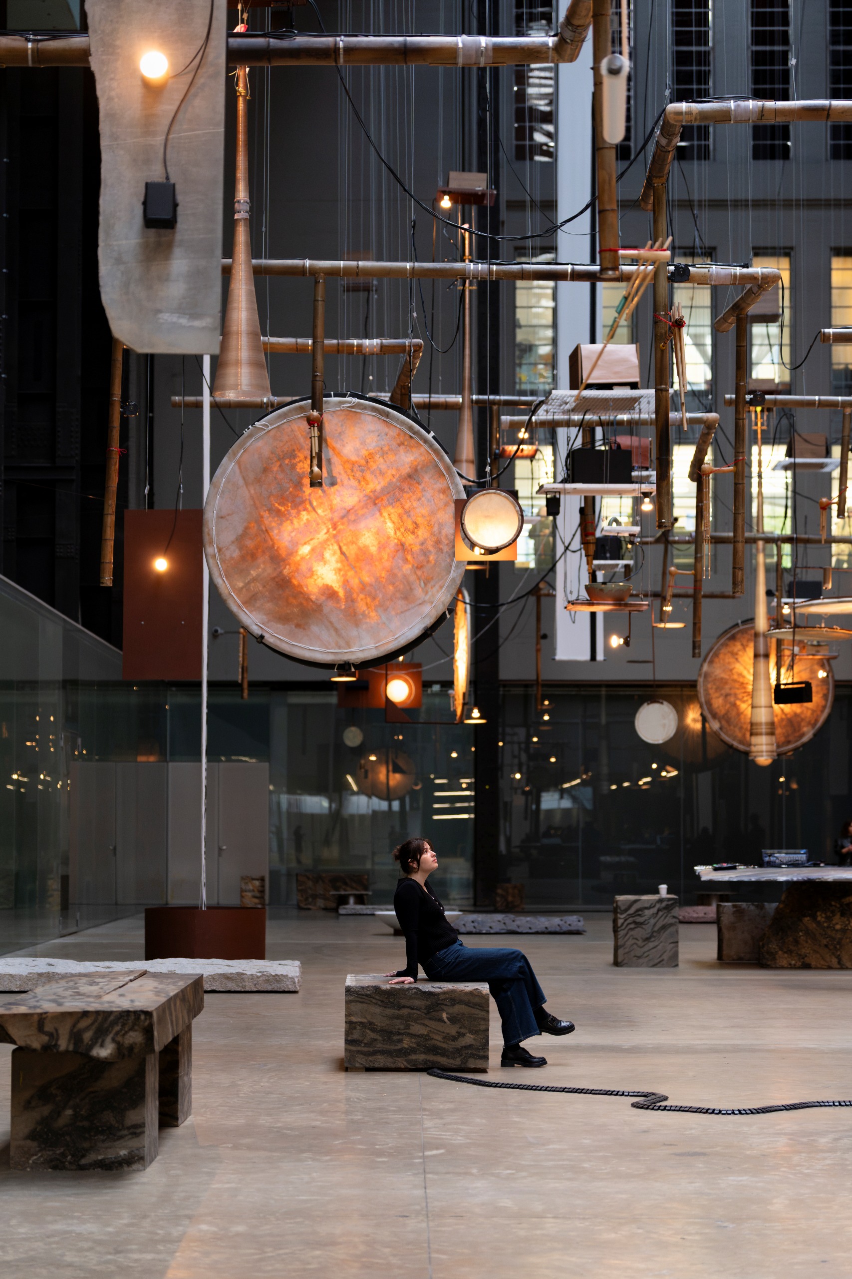 A colossal installation set inside a brutalist building with cement floors and iron and glass walls featured illuminated sculptures in paper and metal, sculptural seats, and bronze piping.
