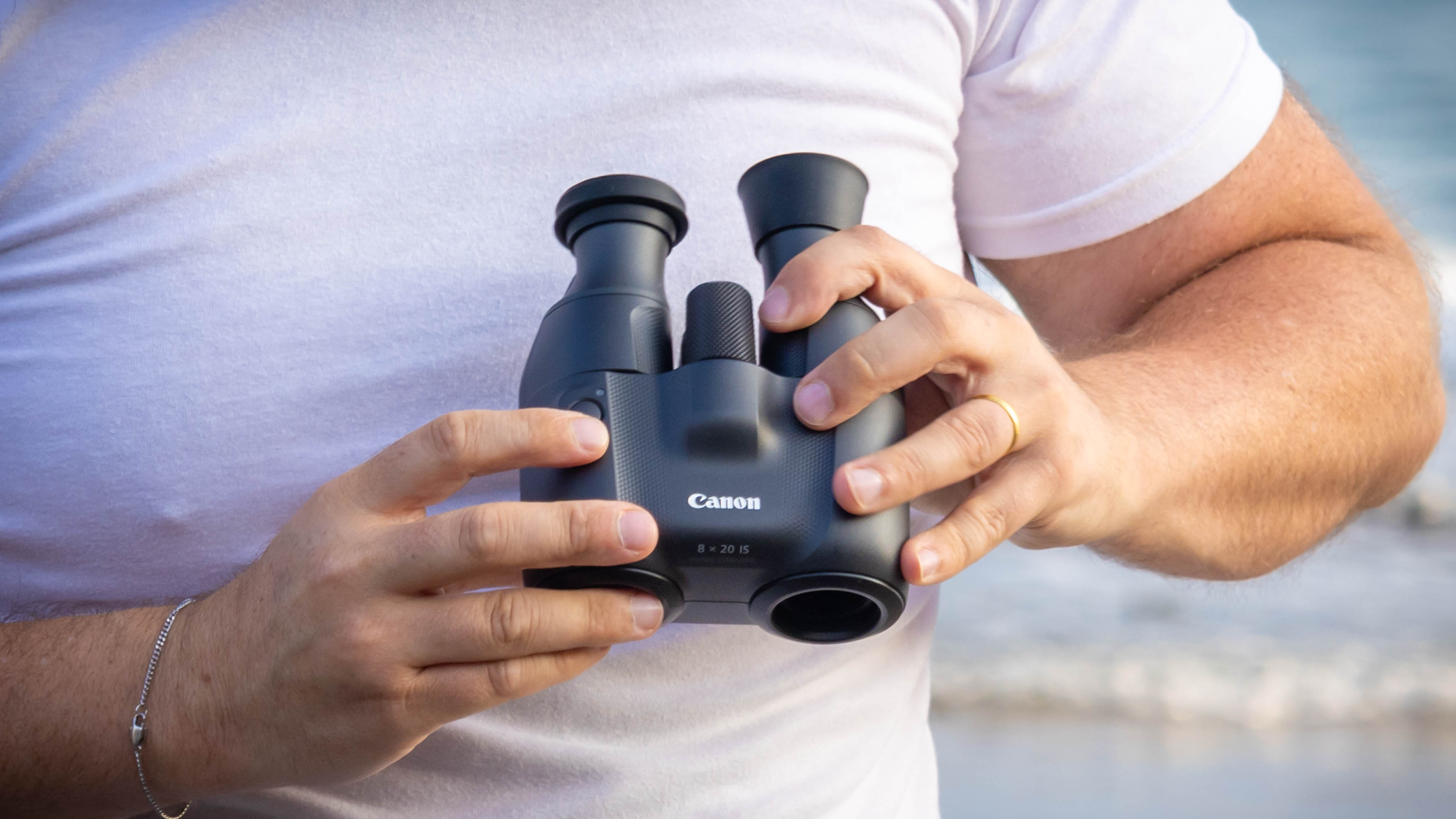 A person holding the Canon 8x20 IS binoculars, with their finger over the IS button.