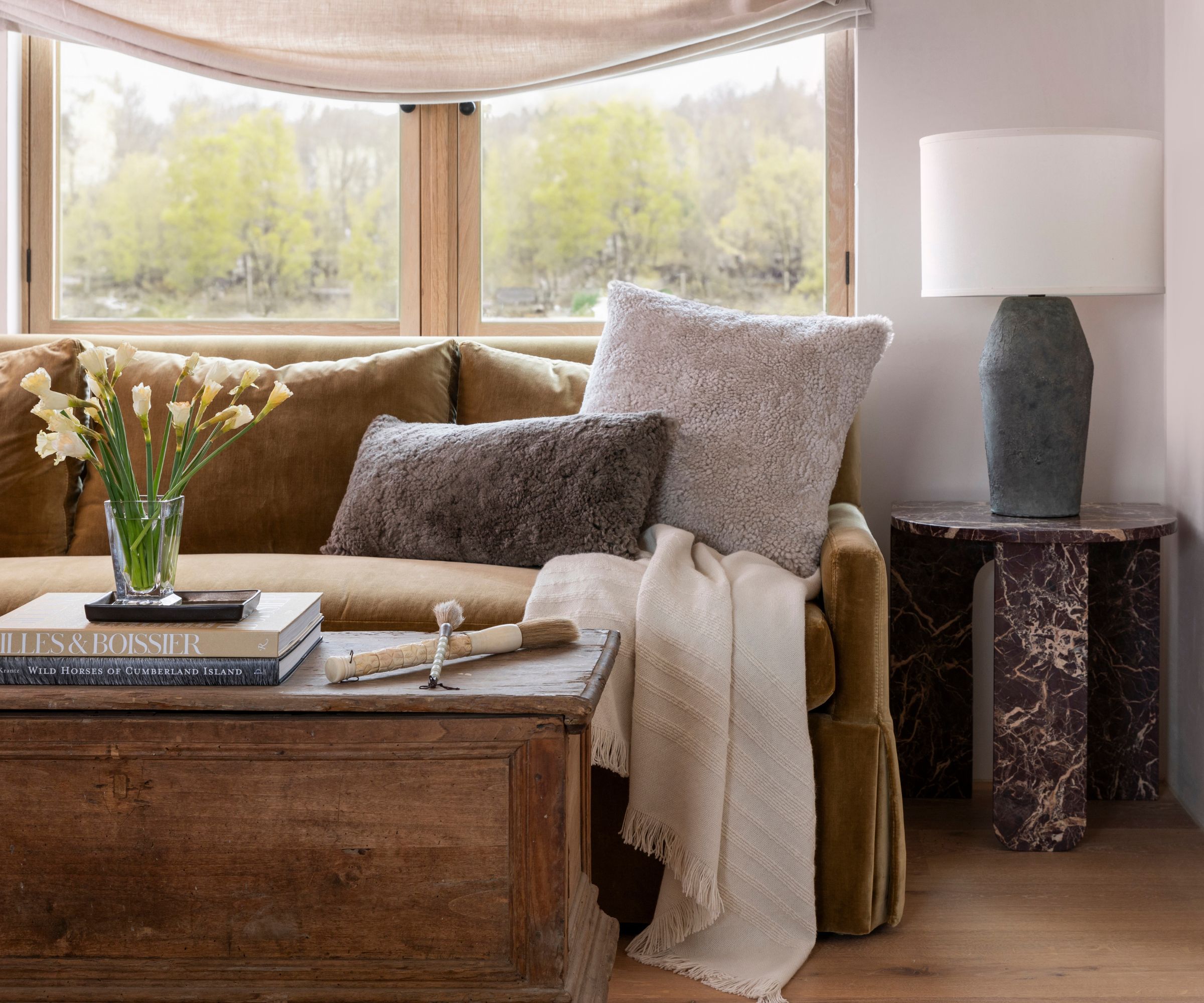 neutral living room with a relaxed roman blind, rustic wooden trunk coffee table, caramel velvet sofa and a burgundy red side table with a lamp