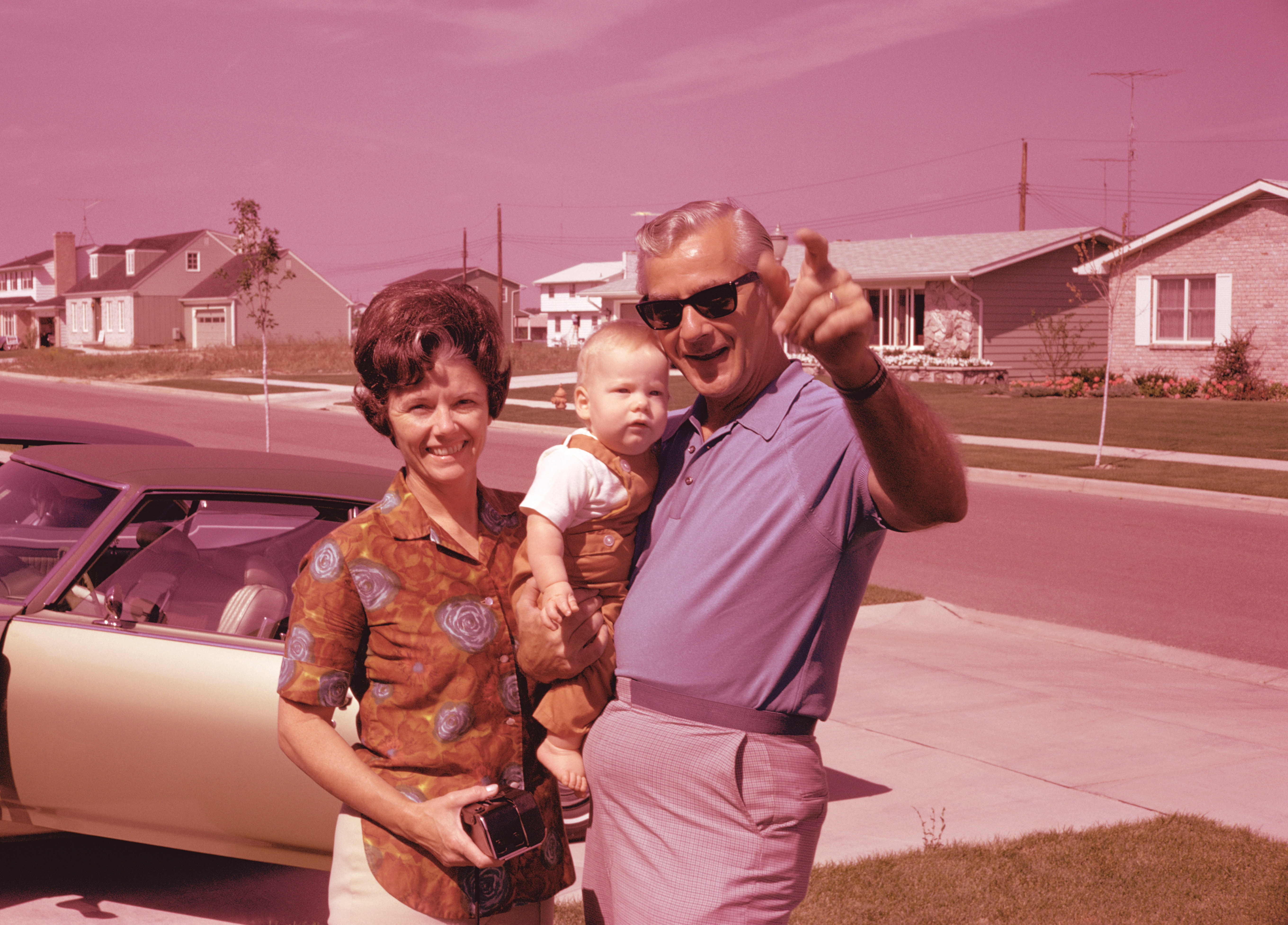 A husband and wife stand on their lawn in front of their car, holding an infant.