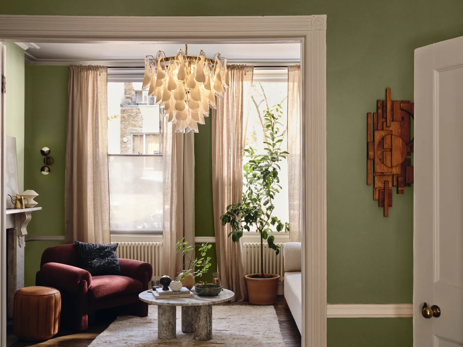 Green and white walls living room with sheer curtains, a red velvet armchair, tan leather ottoman, marble coffee table styled with vases, gray rug, and potted plant