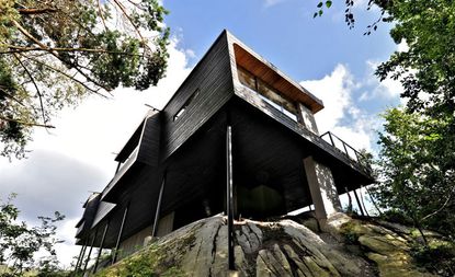 Exterior view from below of the elevated Trekronekabin by Tommie Wilhelmsen - a dark wood house that sits on rocks surrounded by trees under a blue cloudy sky