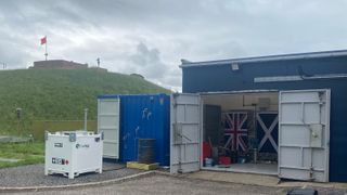 A blue building has its white metal doors wide open, displaying a UK flag, as the building is next to a tall grassy hill under cloudy skies