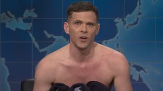 A sheepish Mikey Day sits shirtless on the Weekend Update set for Saturday Night Live.