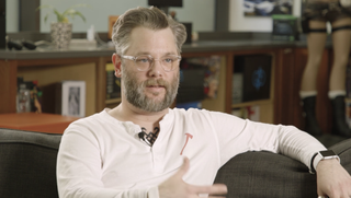 A photo of Cory Barlog sat on a sofa talking to camera