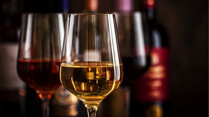 Red, white and rose wine in glasses with a collection of wine bottles in the background.