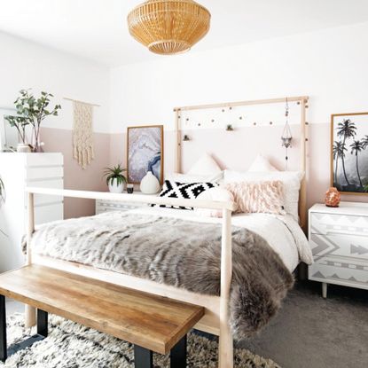 Master teenage girl bedroom with fur throw on double bed, wooden bench and pink and white walls, white and grey chests of drawers as bedside tables. hanging ornaments, fairy lights on the bedhead. 