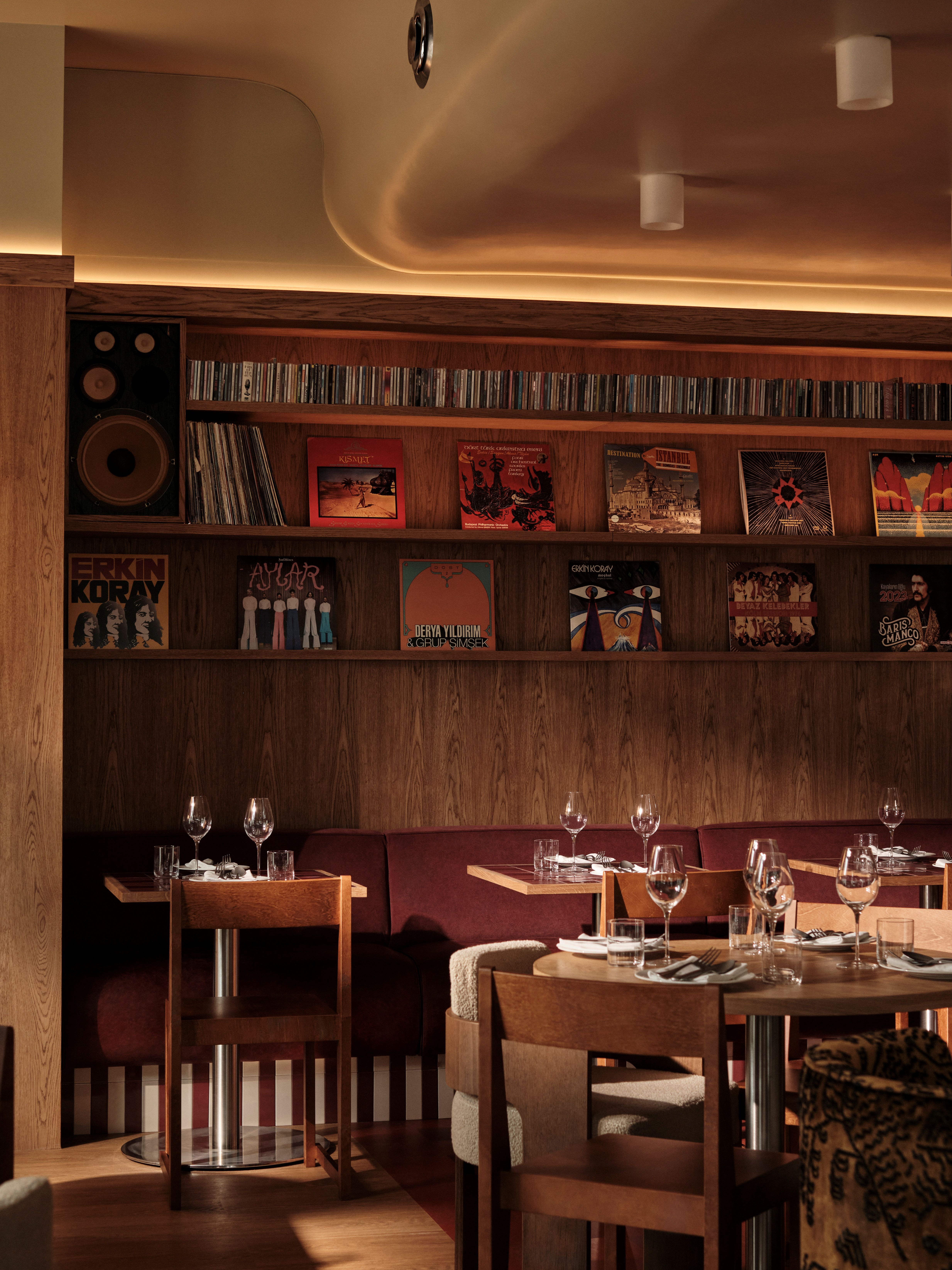 Interior shots of a restaurant decorated with soft wood walls, oversized modular couches in creamy shades, upholstered banquettes in bordeaux and brown shades, sculptural lighting, and a collection of records.