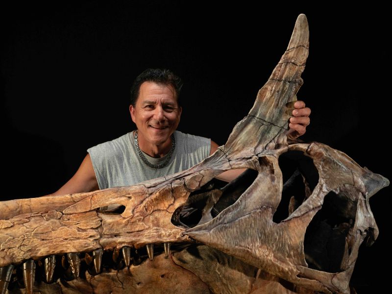 A man with brown hair and a gray tank top stands behind a large brown skull of a dinosaur with a long snout and gray spike on the top of its head.