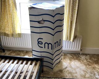Emma mattress packaging box on bedroom carpet next to bed frame