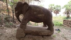 We see the stone-carved statue of an elephant with its trunk curved downward toward the ground. It is in a dirt-covered area of a grass field.